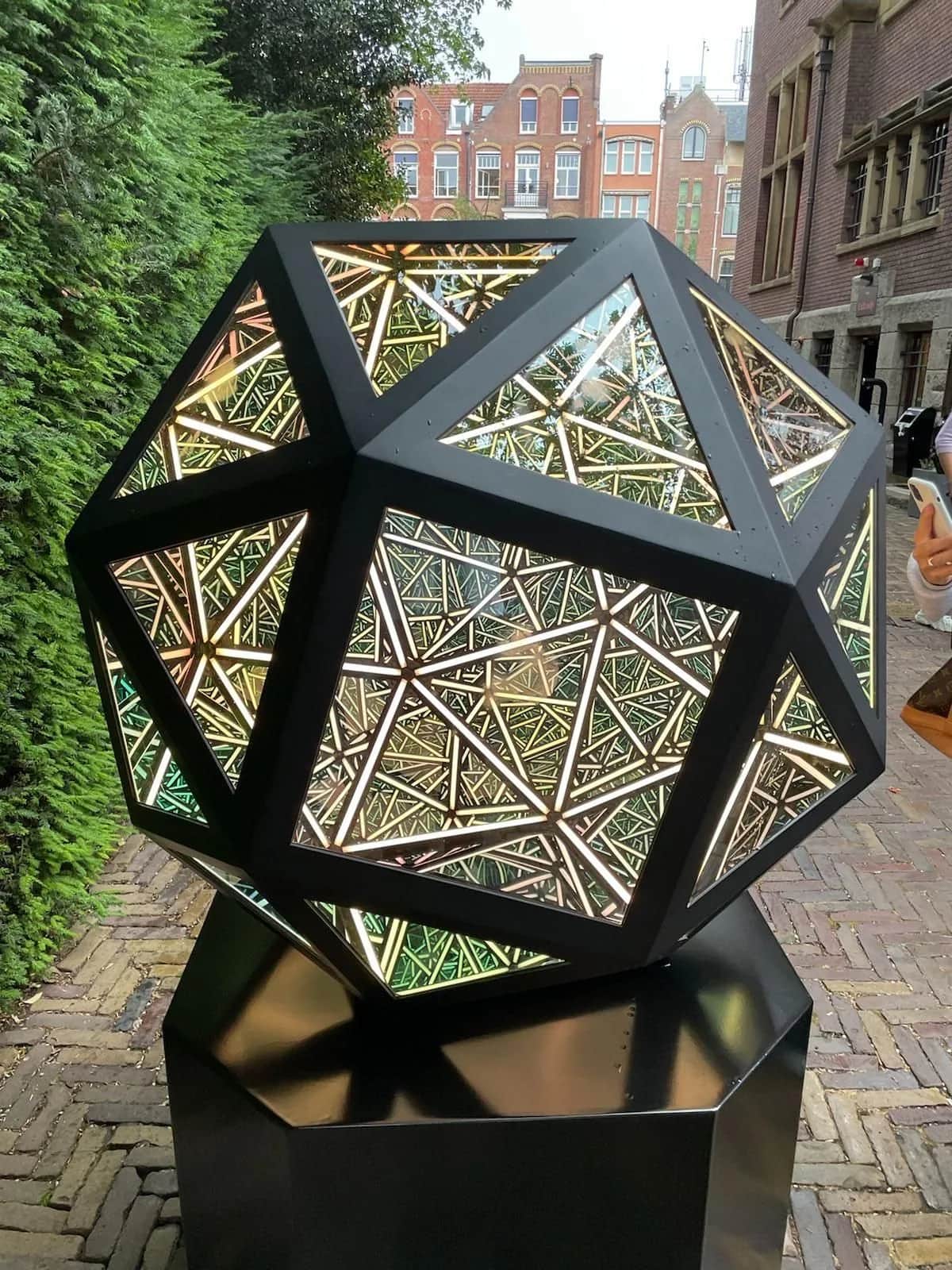 Mirrored geometric sculpture glowing on its plinth on a brick street outside the Moco Museum, Amsterdam 2023