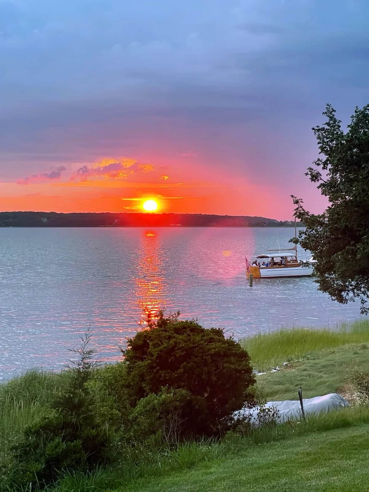 Red sun dropping to the far shore behind a small boat flying a flag, East Hampton 2021