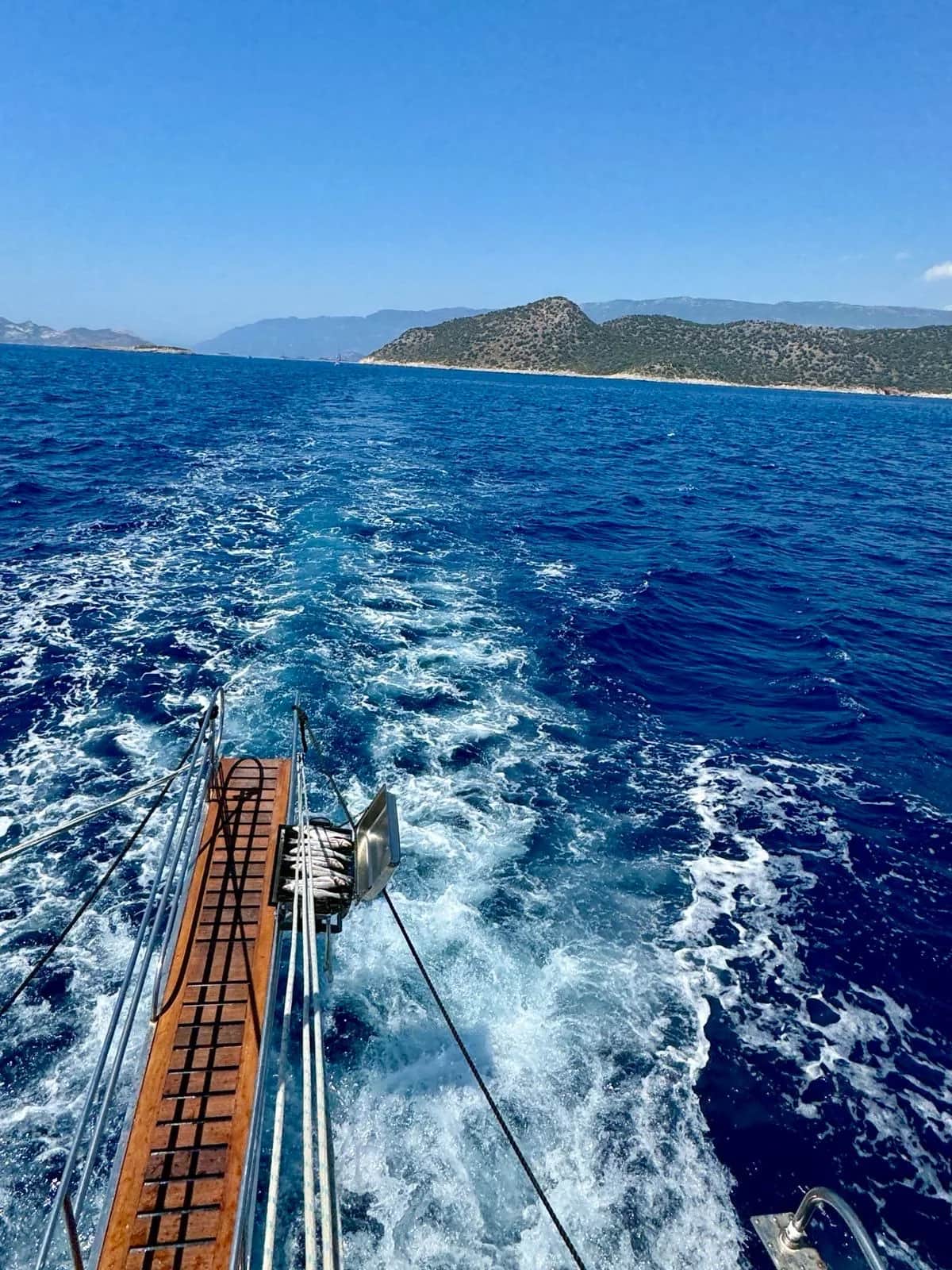 Wake churning behind a wooden boat crossing deep blue water away from a green headland, Kas 2024