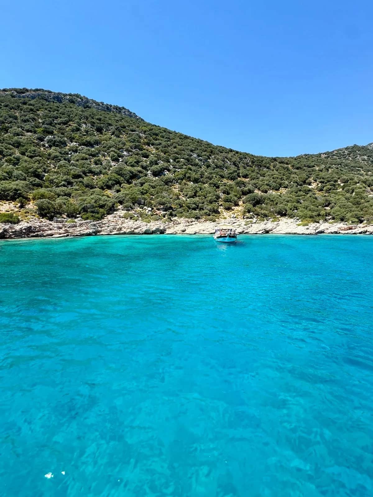 Small boat moored in a clear turquoise cove below a scrubby limestone hillside, Kas 2024