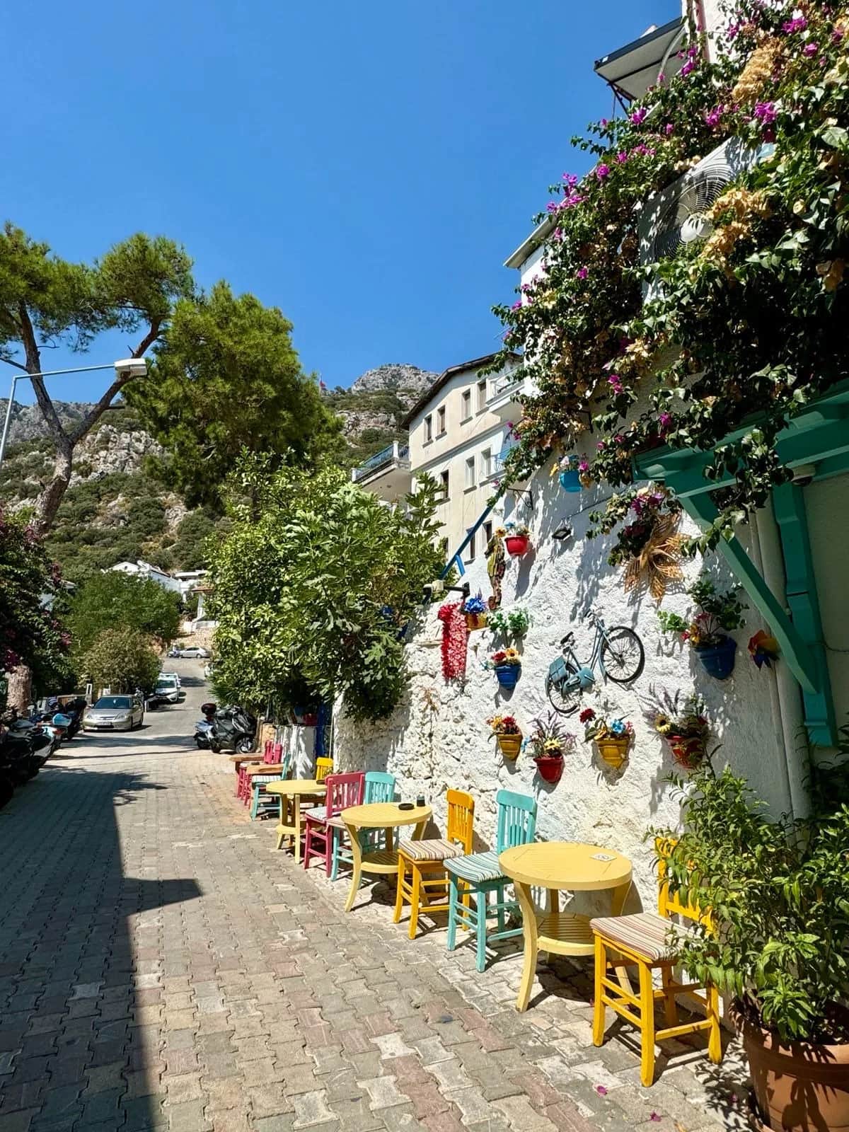 Yellow tables and bright painted chairs along a cobbled lane against a white wall of flower pots, Kas 2024