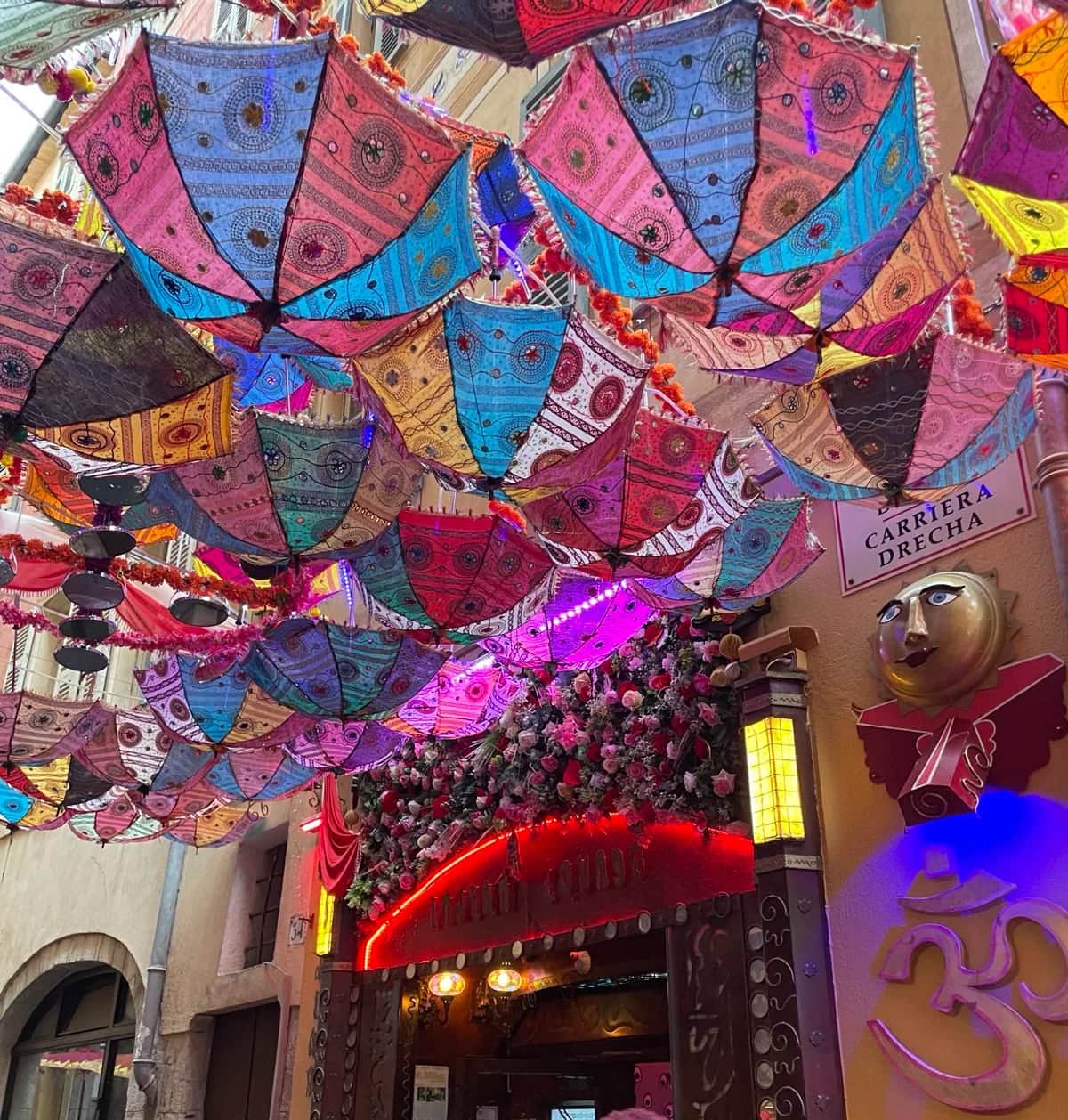 Hundreds of embroidered parasols strung above a narrow old town lane over a glowing shop front, Nice 2023
