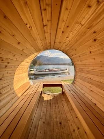 A barrel sauna looking out through its round window to canoes at the lake shore, Jasper